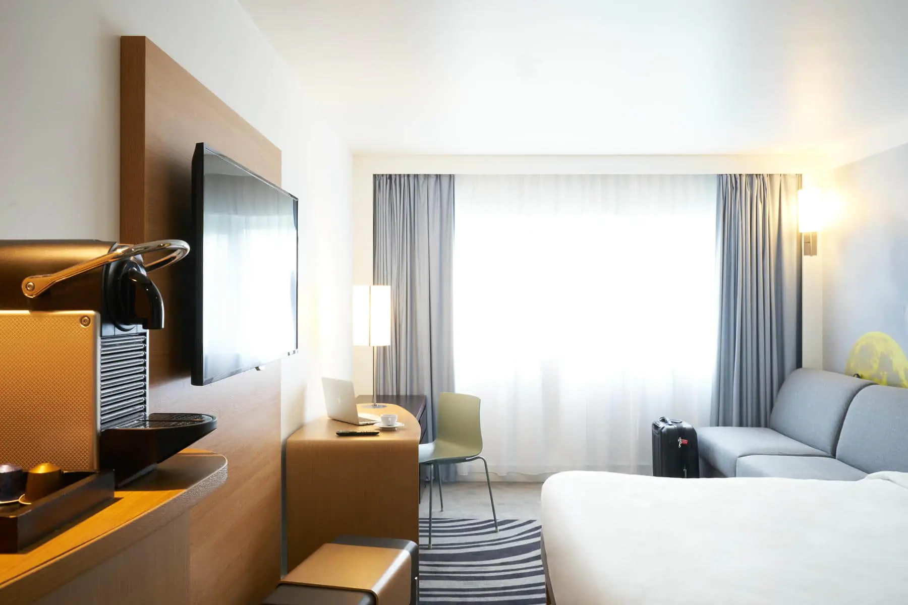 A bright bedroom with a large double bed on the right facing a coffee machine and a wall-mounted flat-screen TV, and at the back of the room a grey sofa on the right, a workspace on the left, and a large window in the background.