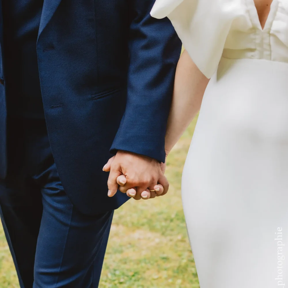 Mariés vus de face, avec un focus sur leurs mains jointes portant des alliances en or, le marié en costume bleu et la mariée en robe blanche décolletée.