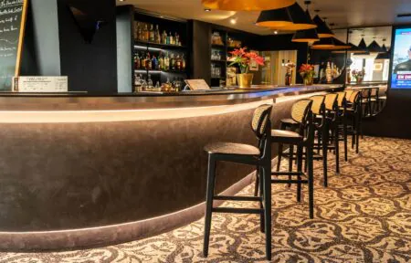 Un bar d'hôtel aux lignes épurées dans les tons chocolat, avec une rangée de tabourets haut noirs et rotin, le bar sur toute la longueur à gauche surmonté de suspensions noir et or, et derrière le bar plusieurs bouteilles exposées.