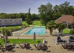 Vue aérienne d'une terrasse d'hôtel avec sol pavé, tables en bois foncé, chaises et parasols fermés au premier plan, une piscine rectangulaire avec chaises longues blanches et palmiers au centre, un bâtiment beige à plusieurs étages à gauche et un bâtiment au toit en tuiles à droite, le tout entouré de pelouses et d'arbres denses, un domaine propice à un séminaire au vert.