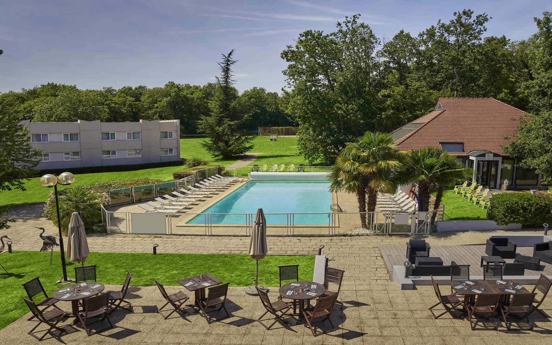 Vue aérienne d'une terrasse d'hôtel avec sol pavé, tables en bois foncé, chaises et parasols fermés au premier plan, une piscine rectangulaire avec chaises longues blanches et palmiers au centre, un bâtiment beige à plusieurs étages à gauche et un bâtiment au toit en tuiles à droite, le tout entouré de pelouses et d'arbres denses, un domaine propice à un séminaire au vert.