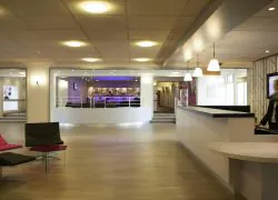 Hall d'hôtel avec sol en bois clair, un fauteuil rose, une chaise noire et une petite table en verre à gauche, un comptoir d'accueil blanc incurvé avec un membre du personnel à droite sous des suspensions lumineuses, et un espace bar éclairé en violet visible derrière une estrade en arrière-plan, le point d'accueil d'un hôtel pour séminaire.