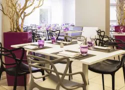 Intérieur de restaurant d'hôtel avec sol carrelé beige, tables blanches dressées avec sets de table noirs, serviettes pliées, verres à vin transparents et gobelets violets au premier plan, chaises noires et grises au dossier incurvé autour de chaque table, et des compositions de branches dans des bacs magenta de part et d'autre, avec d'autres tables visibles à travers des baies vitrées donnant sur une terrasse en arrière-plan, un cadre pour la restauration sur place.