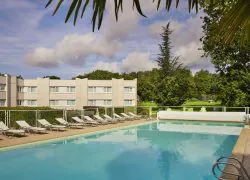 Piscine extérieure à l'eau turquoise avec échelle métallique au premier plan, chaises longues blanches alignées sur une zone pavée derrière une clôture de sécurité vitrée à gauche, et un bâtiment d'hôtel beige à plusieurs étages bordé d'une pelouse et d'arbres en arrière-plan, encadré par des feuilles de palmier en hauteur, au sein d'un domaine propice à un séminaire bien-être.
