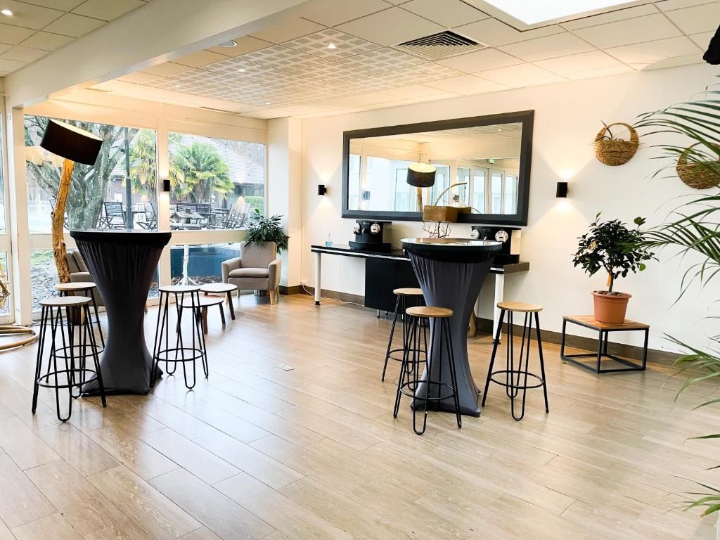 Espace lounge intérieur avec sol en bois clair, deux mange-debout drapés de noir entourés de tabourets de bar en bois aux pieds noirs en épingle au premier plan, un bar à café noir et un grand miroir contre le mur du fond, et un fauteuil beige, des plantes en pot et de grandes fenêtres donnant sur une terrasse, parfait pour un afterwork.