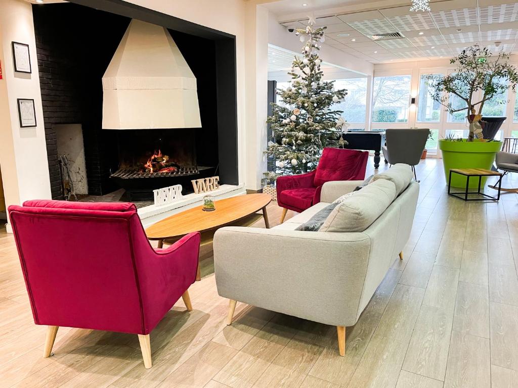 Salon d'accueil d'hôtel avec sol en bois clair, une cheminée noire avec bûches allumées et hotte blanche à gauche, deux fauteuils en velours bordeaux et un canapé beige autour d'une table basse ovale en bois au premier plan, et un sapin de Noël décoré près de grandes fenêtres à droite, un cadre chaleureux pour de l'hébergement groupe.