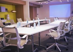 Salle de réunion avec moquette grise, tables blanches disposées en U avec chaises de bureau à dossier résille sur roulettes, bouteilles d'eau et blocs-notes placés à chaque place, et un grand écran de projection lumineux sur le mur du fond, aménagée en salle de formation.