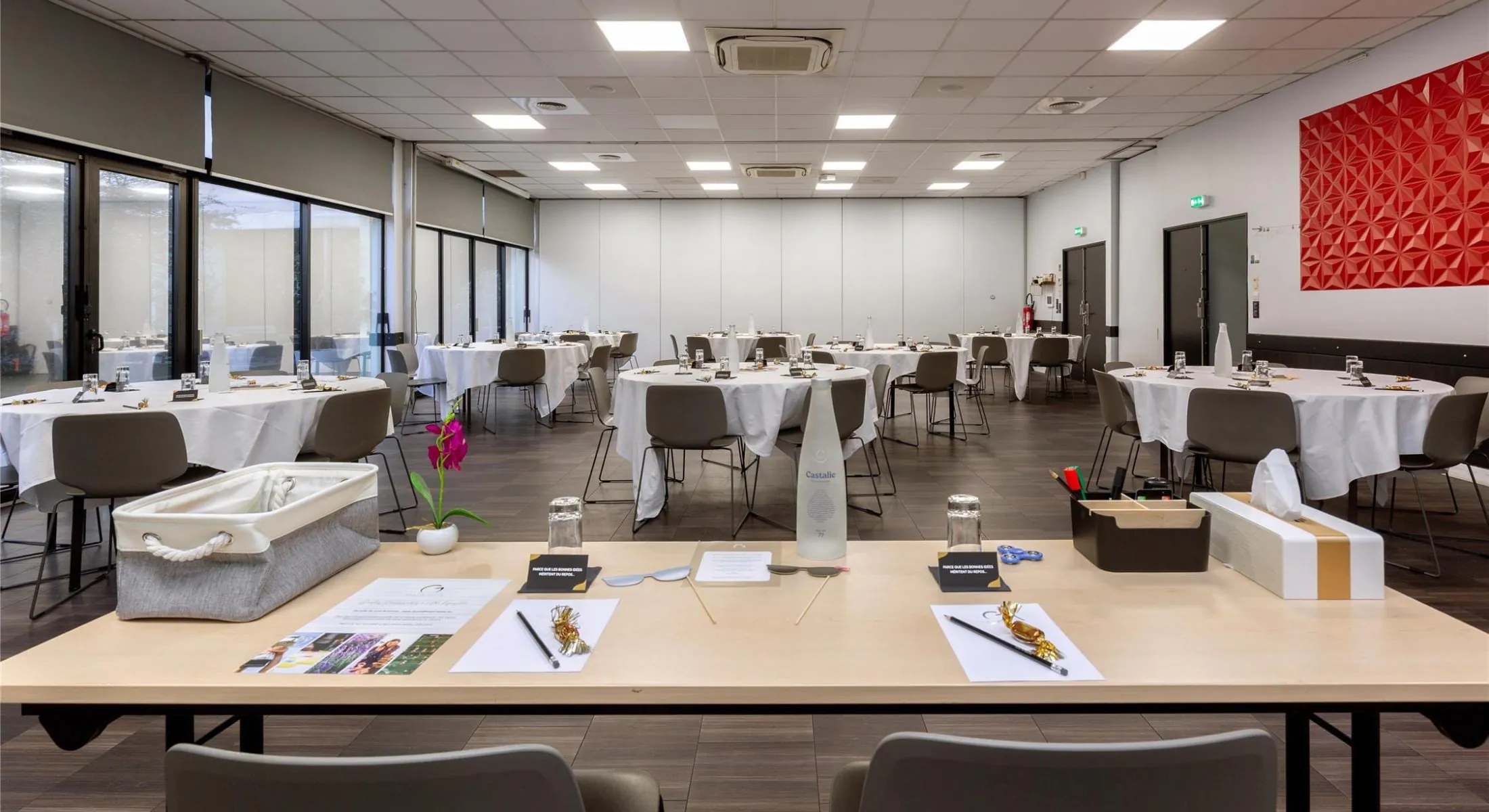 Large hotel meeting room rental with a light wood table set for a seminar in the foreground featuring a Castalie water bottle, notepads, pencils, a grey fabric basket and a pink orchid, multiple round white tablecloth tables with grey chairs arranged throughout the room, a large red geometric relief panel on the right white wall, grey roller blinds over wide bay windows on the left, and a white operable partition wall at the back.