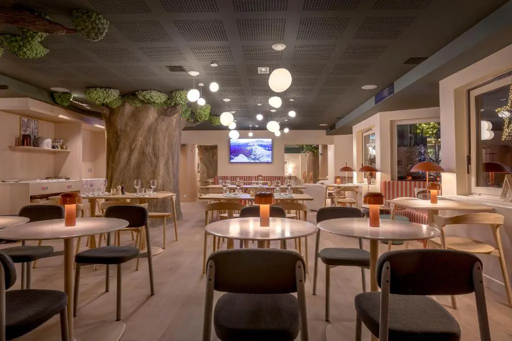 Restaurant d'hôtel avec plafond sombre perforé orné de suspensions globes, comprenant un grand tronc d'arbre décoratif avec feuillage vert à gauche, des tables rondes avec chaises sombres et en bois clair et lampes de table cuivrées, un téléviseur mural et des banquettes rayées rouge et blanc en arrière-plan, et un sol en bois clair, pour une restauration sur place.