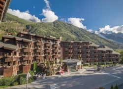 Vue extérieure d'un grand complexe hôtelier de montagne aux façades de chalet en bois sombre avec de nombreux balcons fleuris, comprenant une rotonde d'entrée vitrée au centre, bordant une route avec des sommets alpins enneigés et une forêt de pins en toile de fond, pour un hébergement groupe grande capacité et des séminaires à la montagne.
