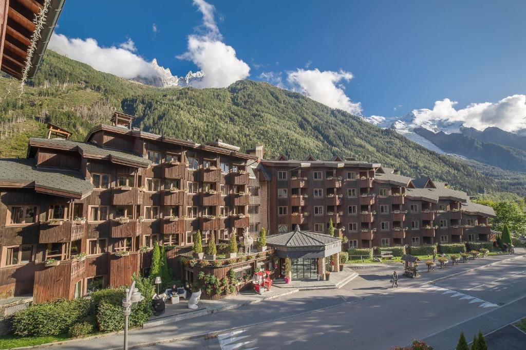 Vue extérieure d'un grand complexe hôtelier de montagne aux façades de chalet en bois sombre avec de nombreux balcons fleuris, comprenant une rotonde d'entrée vitrée au centre, bordant une route avec des sommets alpins enneigés et une forêt de pins en toile de fond, pour un hébergement groupe grande capacité et des séminaires à la montagne.