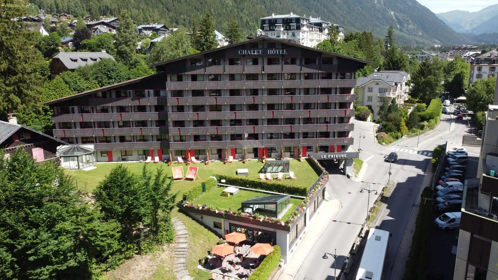 Vue aérienne d’un hôtel de séminaire montagne, avec au centre un vaste bâtiment de style chalet doté de nombreux balcons donnant sur des espaces verts aménagés et des zones de détente extérieures ; entouré d’arbres, de bâtiments voisins et de routes, l’établissement est niché au cœur d’une vallée de montagne.
