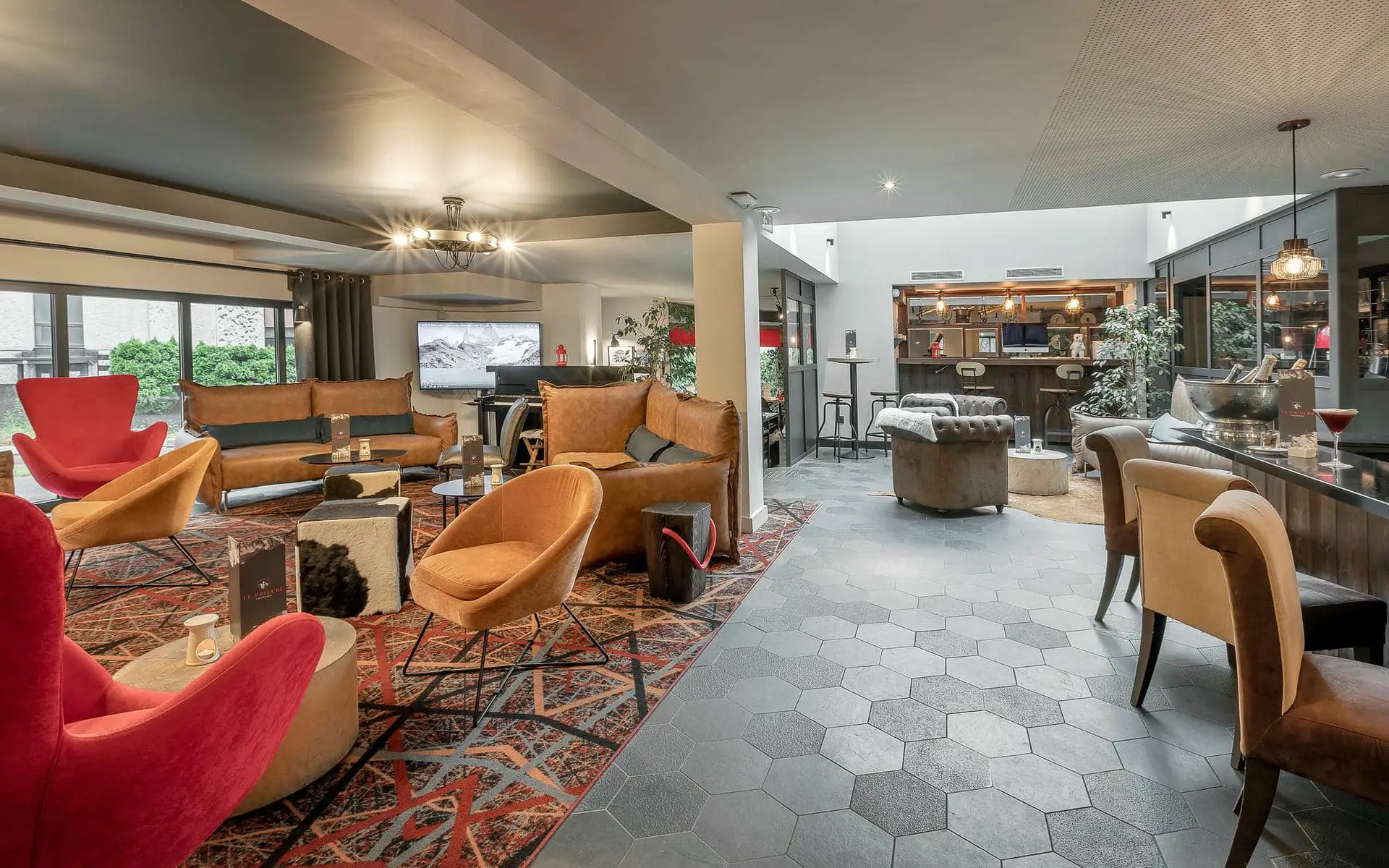 Espace afterwork au sol en carreaux hexagonaux foncés, avec canapés en cuir et fauteuils colorés disposés autour de tables basses au premier plan et au centre, tapis à motifs délimitant l’espace salon à gauche, et espace bar avec tabourets hauts à l’arrière-plan ; l’éclairage chaleureux, le mobilier contemporain et les plantes décoratives créent une ambiance conviviale et détendue.