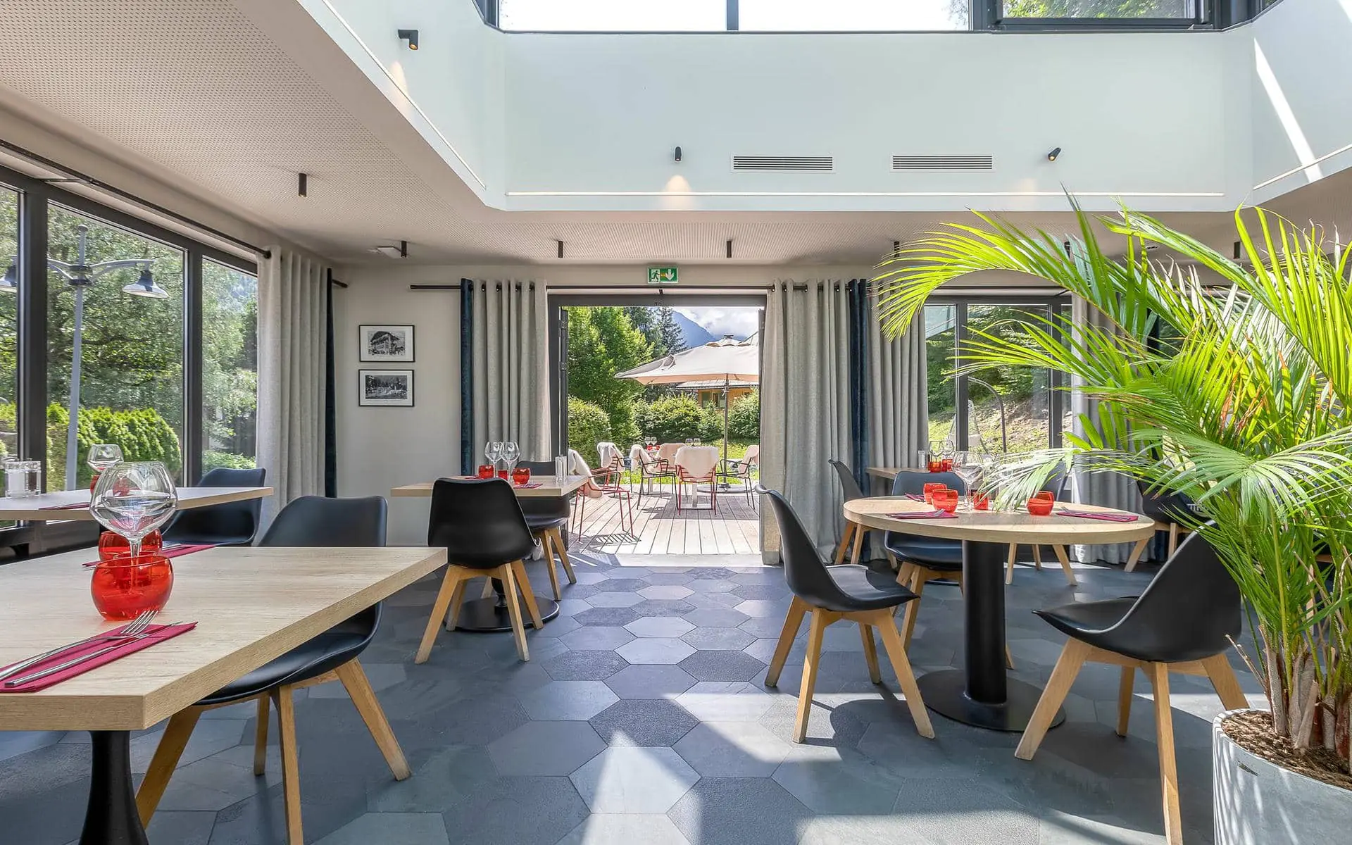 Espace de restauration sur place au sol en carreaux hexagonaux foncés, avec tables dressées agrémentées de photophores rouges et chaises noires réparties au premier plan et au centre, grandes baies vitrées toute hauteur de part et d’autre, et ouverture sur une terrasse ensoleillée à l’arrière-plan ; un grand palmier en pot à droite et une généreuse lumière naturelle créent une ambiance contemporaine.