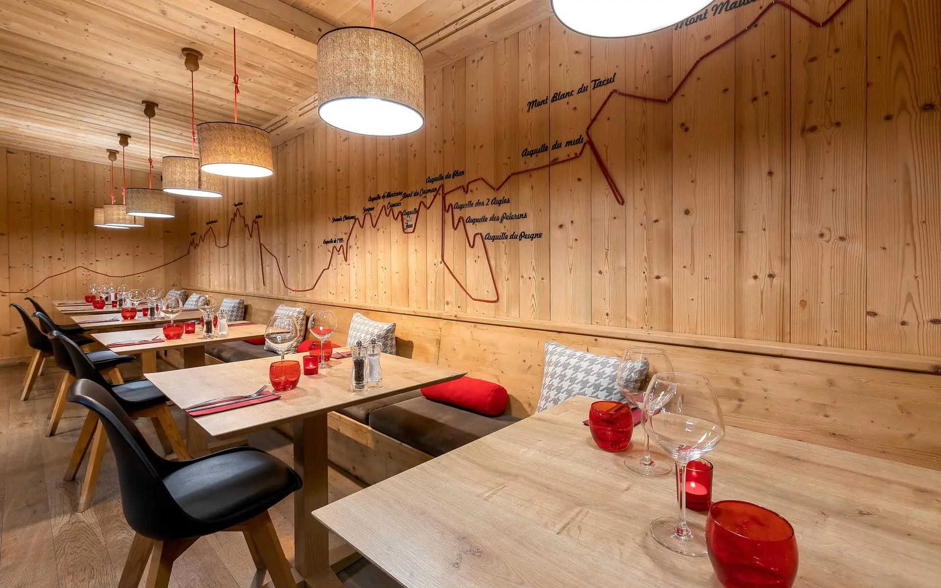 Espace de restauration sur place, avec à gauche et au centre des tables en bois dressées avec verrerie, photophores rouges et chaises noires, tandis qu’une banquette continue agrémentée de coussins longe le mur de droite ; un habillage en bois décoré d’un tracé de montagne et des suspensions lumineuses créent une ambiance chaleureuse d’inspiration alpine.