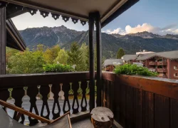 Vue depuis un balcon d’hébergement de groupe pour un séminaire à la montagne, avec garde-corps en bois et chaises longues au premier plan, surplombant des bâtiments hôteliers de style chalet à droite et des sommets montagneux boisés s’étendant à l’arrière-plan.