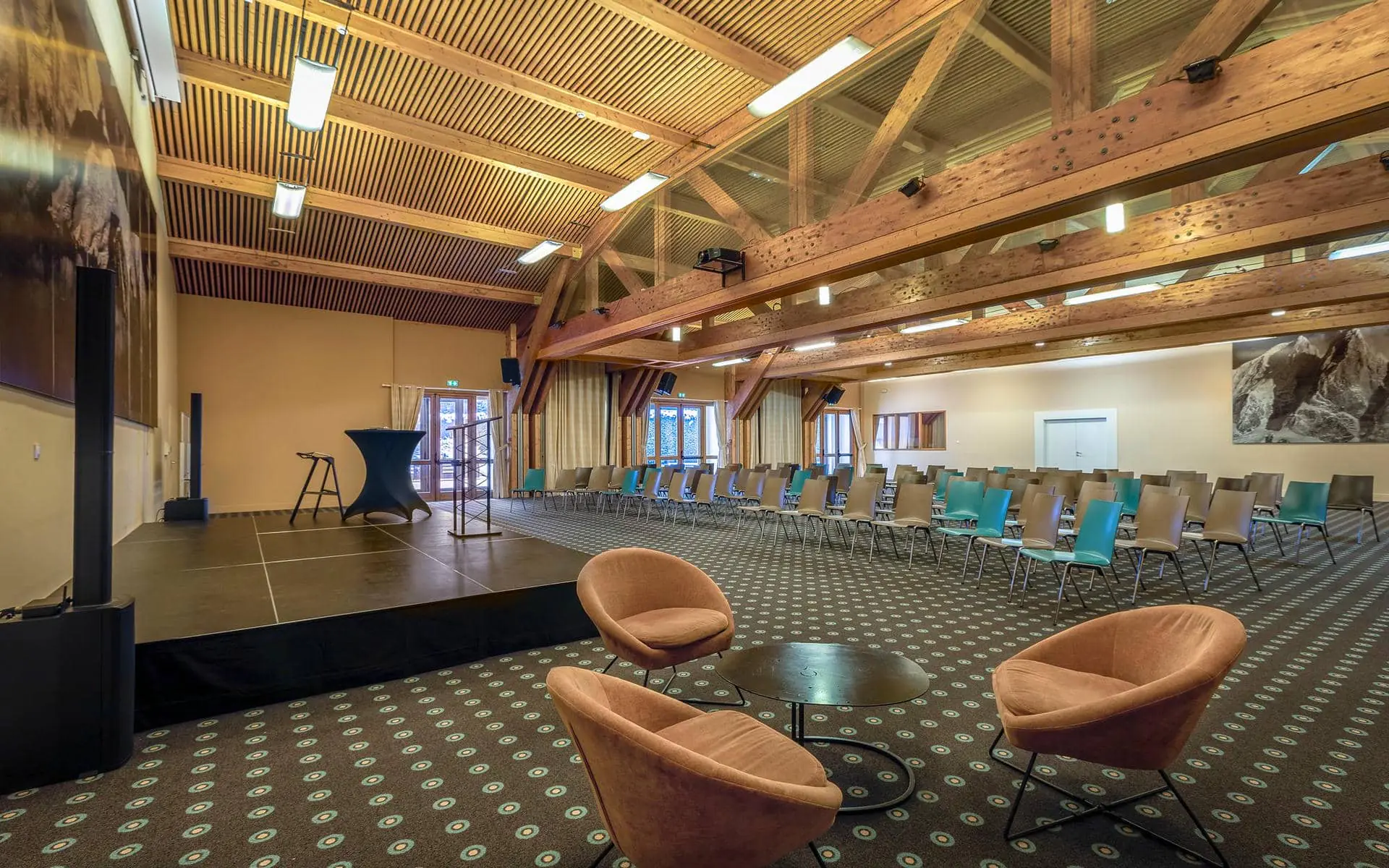 Salle de conférence proposée en location de salle, avec à gauche une scène équipée d’un pupitre et de matériel audiovisuel, tandis que des rangées de chaises sont disposées face à l’espace de présentation sous une charpente apparente en bois ; au premier plan, un coin salon composé de fauteuils et d’une table basse complète l’ensemble.