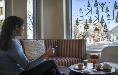 Une femme tenant une tasse bleue est assise sur un canapé rayé rouge et blanc avec coussin en maille blanche, face à une grande fenêtre décorée du logo de l'hôtel Lykke et de sapins verts, avec une petite table ronde portant théière et pâtisseries au premier plan et une rue de village de montagne enneigée visible à l'extérieur, une scène de séjour pour un séminaire à la montagne.