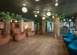 Réception insolite d’hôtel au sol en bois, avec colonnes décoratives en forme d’arbres réparties dans l’espace, fauteuils sculpturaux rayés à gauche et fauteuils lounge bleus à droite ; un comptoir de réception se trouve à l’arrière-plan sous des suspensions sphériques.