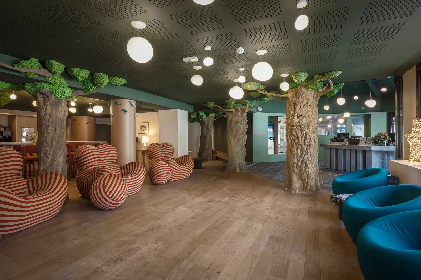 Réception insolite d’hôtel au sol en bois, avec colonnes décoratives en forme d’arbres réparties dans l’espace, fauteuils sculpturaux rayés à gauche et fauteuils lounge bleus à droite ; un comptoir de réception se trouve à l’arrière-plan sous des suspensions sphériques.