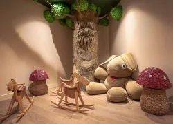Espace jeux enfants d'hôtel avec sol en bois clair, comprenant un grand tronc d'arbre décoratif avec visage sculpté et feuillage vert dans le coin, deux chevaux à bascule en bois, un grand lapin en peluche et deux poufs en osier rouge en forme de champignon disposés autour.
