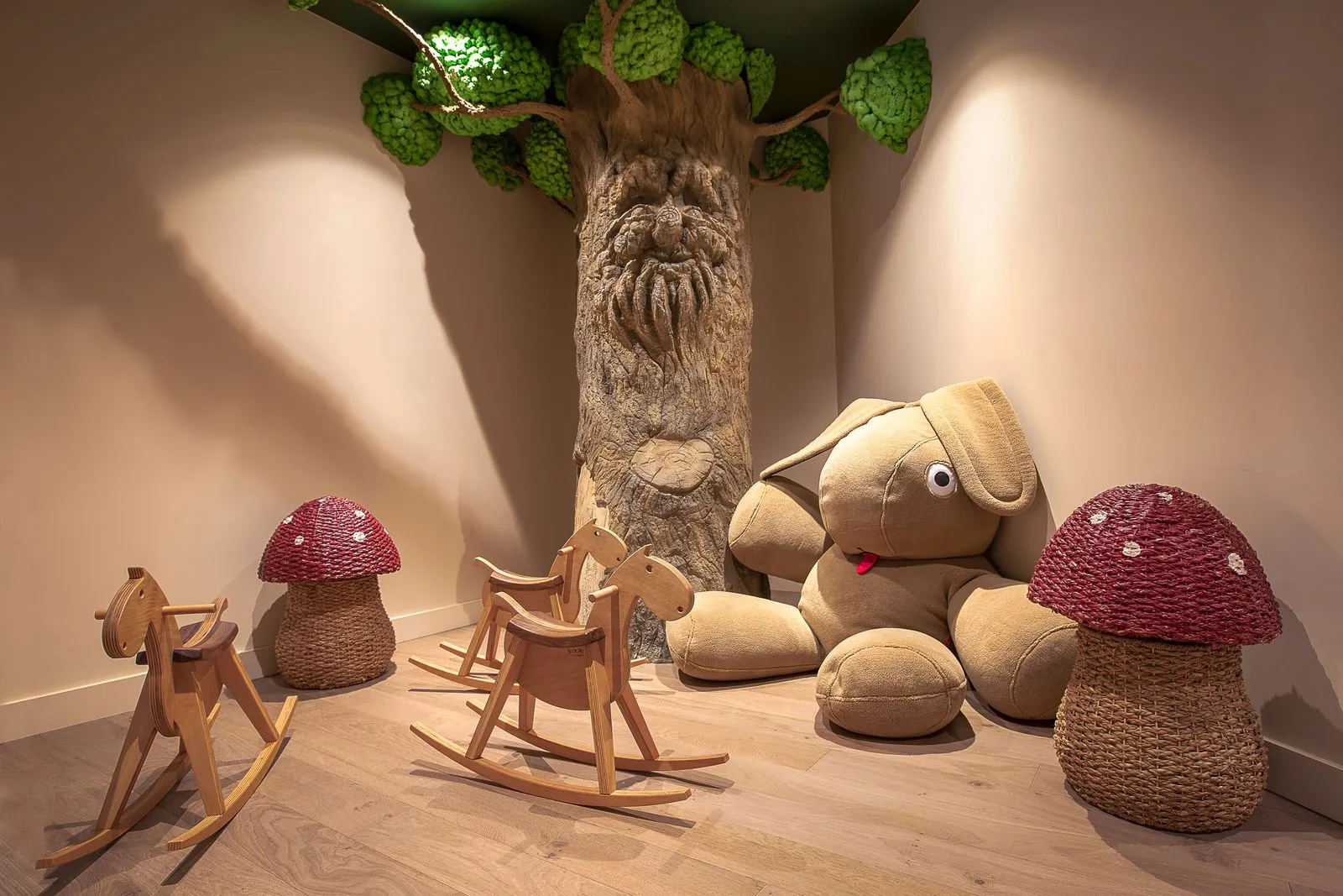 Espace jeux enfants d'hôtel avec sol en bois clair, comprenant un grand tronc d'arbre décoratif avec visage sculpté et feuillage vert dans le coin, deux chevaux à bascule en bois, un grand lapin en peluche et deux poufs en osier rouge en forme de champignon disposés autour.
