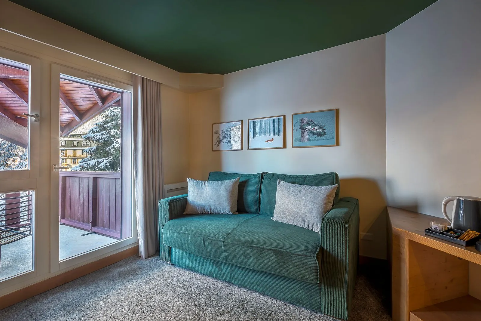 Chambre d’hôtel pour un séminaire à la montagne, avec au centre un canapé vert agrémenté de coussins clairs sous des illustrations inspirées de la nature, tandis qu’à gauche une grande porte vitrée s’ouvre sur un balcon avec vue sur les montagnes ; à droite, un meuble d’accueil en bois équipé d’une bouilloire complète le plafond vert foncé et la moquette claire.