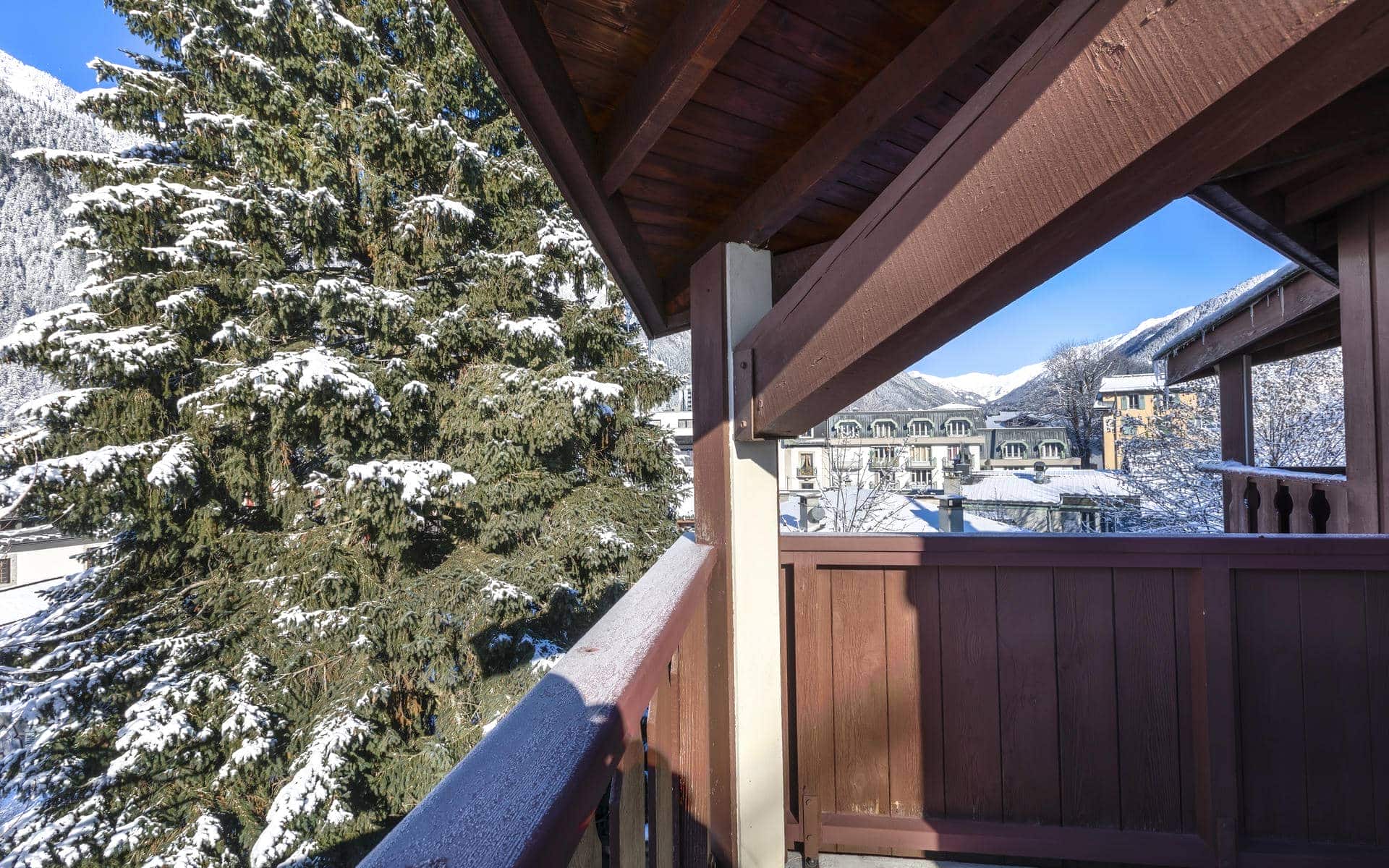Vue depuis un balcon couvert lors d’un séminaire à la montagne, avec au premier plan à gauche des sapins enneigés, des garde-corps en bois encadrant la scène à droite, et en arrière-plan des bâtiments hôteliers ainsi que des sommets enneigés sous un ciel bleu dégagé.