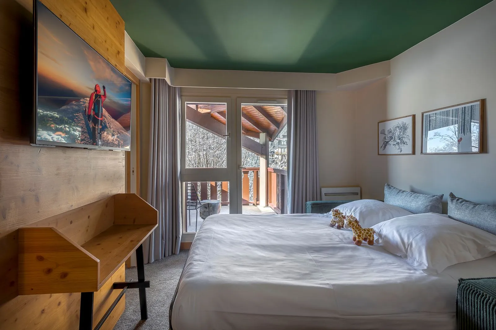 Chambre d’hôtel adaptée à l’hébergement de groupe, avec au premier plan un lit double habillé de linge blanc face à une baie vitrée ouvrant sur un balcon, à gauche un bureau en bois surmonté d’un téléviseur mural et, à droite, un canapé vert sous des illustrations encadrées, dans une décoration mêlant bois clair, murs blancs et plafond vert foncé.