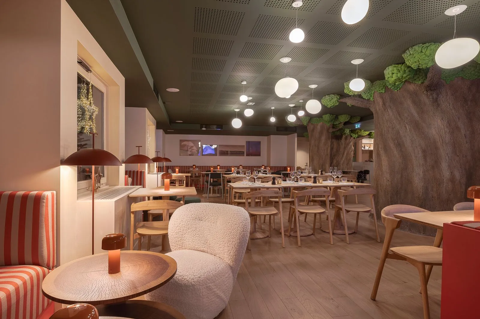 Restaurant d'hôtel avec plafond vert foncé orné de suspensions globes, comprenant un grand tronc d'arbre décoratif sur le mur de droite, des tables et chaises en bois clair au centre, un canapé rayé rouge et blanc avec fauteuil bouclette et lampe cuivrée au premier plan, et un écran télévision visible en arrière-plan, une salle insolite pour prendre ses repas en séminaire.