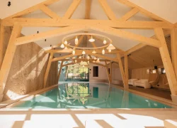 Espace piscine intérieure pour un séminaire bien-être, avec long bassin aux reflets turquoise au centre sous une charpente apparente en bois et des suspensions lumineuses, un mur en pierre texturée à gauche et des assises de détente, dont des fauteuils suspendus, à droite.