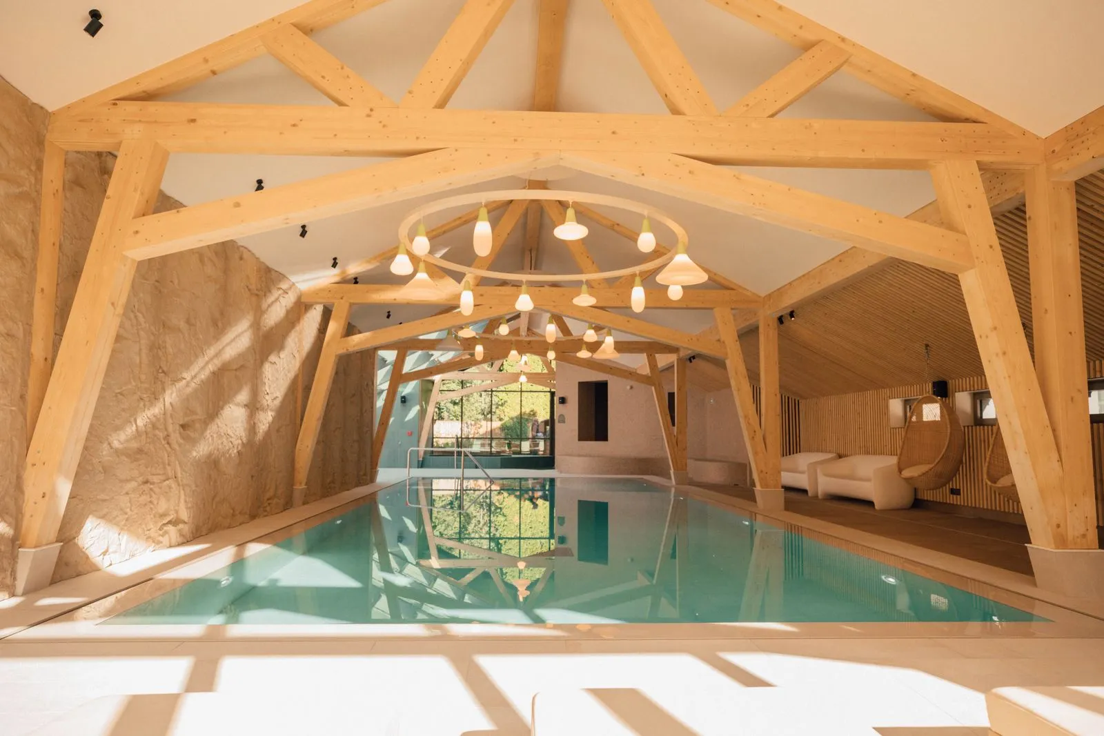 Espace piscine intérieure pour un séminaire bien-être, avec long bassin aux reflets turquoise au centre sous une charpente apparente en bois et des suspensions lumineuses, un mur en pierre texturée à gauche et des assises de détente, dont des fauteuils suspendus, à droite.
