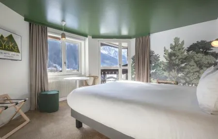 Chambre d’hôtel pour un séminaire à la montagne, avec au premier plan un grand lit double habillé de linge blanc, tandis qu’à gauche une petite table ronde, un tabouret et un porte-bagages sont disposés près de deux fenêtres encadrées de rideaux taupe ; en fond, une porte vitrée s’ouvre sur un balcon avec vue sur les montagnes, et une fresque murale inspirée de la forêt complète le plafond vert foncé et la moquette claire.