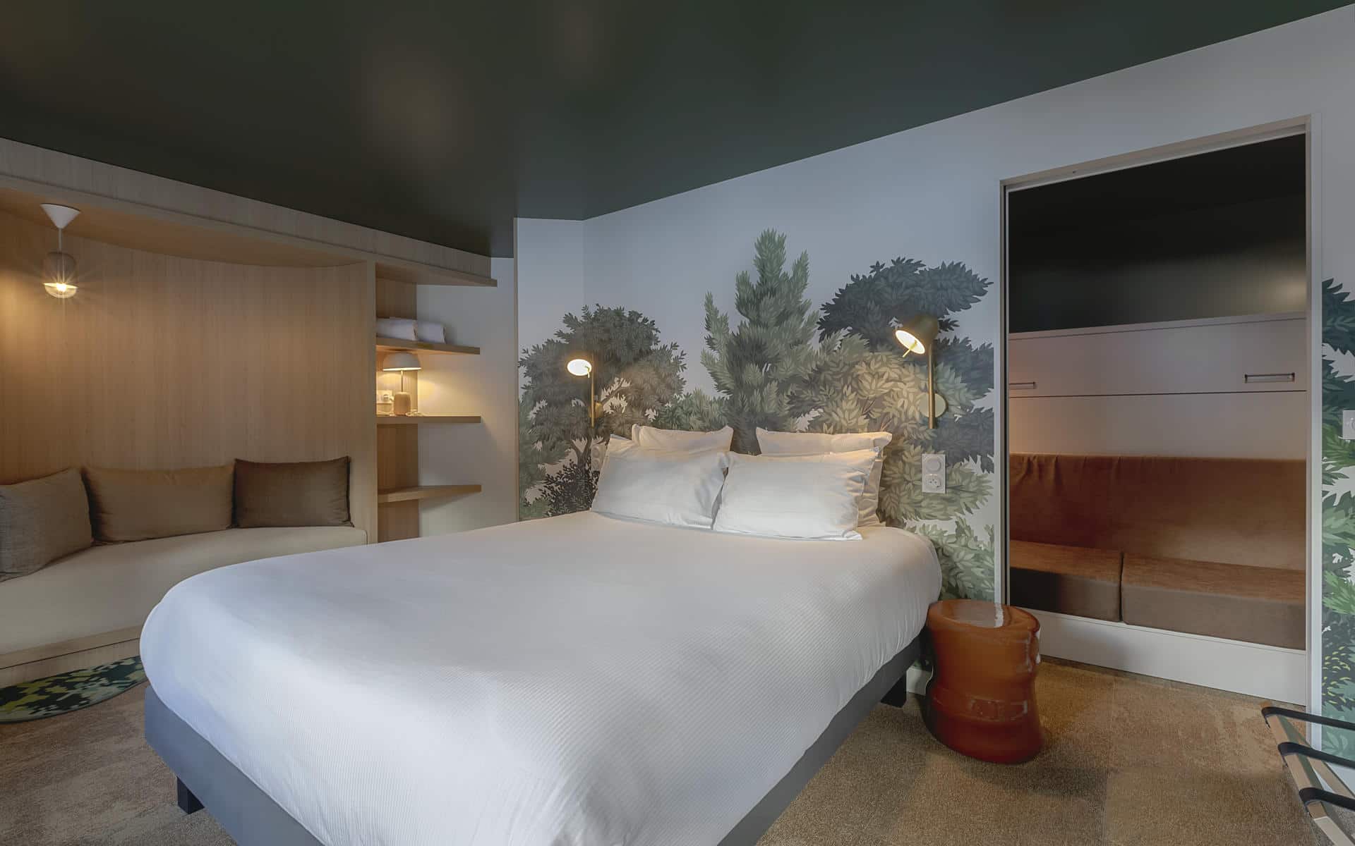 Chambre d’hôtel pour un séminaire à la montagne, avec au centre un grand lit double habillé de linge blanc adossé à une fresque murale inspirée de la forêt, tandis qu’à gauche une alcôve aménagée avec banquette à coussins et étagères en bois crée un coin détente ; à droite, un espace de rangement ouvert avec banquette prolonge l’espace nuit, complété par un plafond vert foncé et une moquette claire.