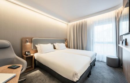 Hotel twin room with grey carpet and warm LED ceiling lighting, featuring two single beds with a shared grey padded headboard framed in light wood and reading sconces, a wooden bedside table with a telephone, a grey ergonomic armchair on the left, a large window with beige curtains and a grey round pouf on the right.
