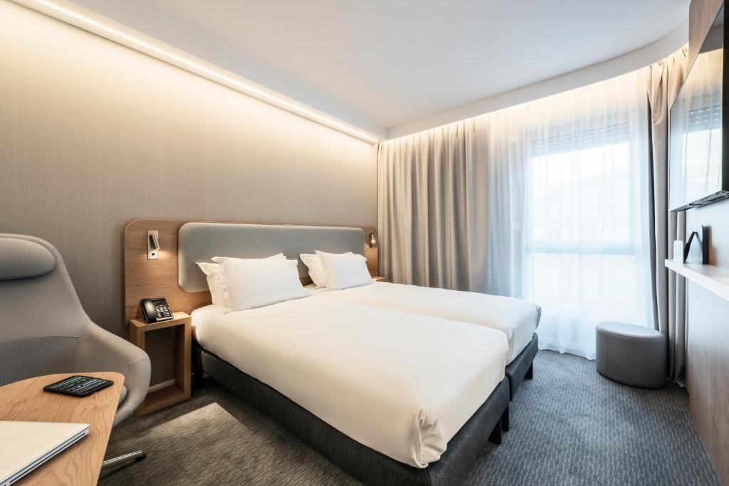 Hotel twin room with grey carpet and warm LED ceiling lighting, featuring two single beds with a shared grey padded headboard framed in light wood and reading sconces, a wooden bedside table with a telephone, a grey ergonomic armchair on the left, a large window with beige curtains and a grey round pouf on the right.