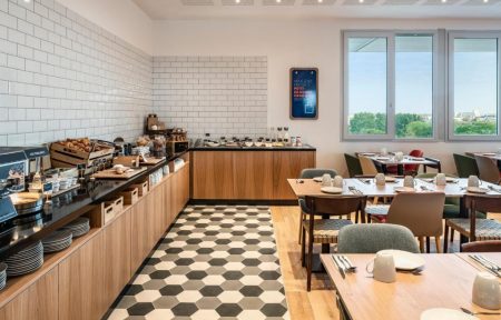 Hotel breakfast room with a black and white hexagonal tiled floor, featuring a wooden buffet counter with pastries, cereals and coffee cups along a white subway tile wall on the left, and wooden dining tables with mixed grey, green and brown chairs on a light wood floor on the right, with large windows overlooking greenery in the background.