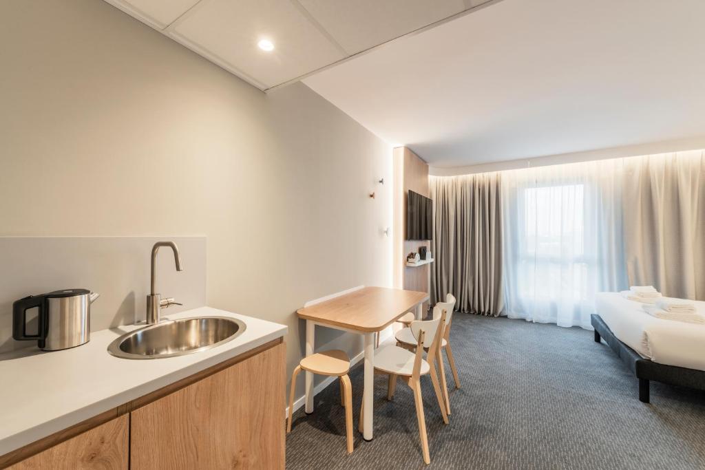 Hotel room with grey carpet and white walls, featuring a light wood kitchenette with a round sink and kettle on the left, a small wooden dining table with chairs in the centre, and a double bed with grey curtains and a wall-mounted television in the background.