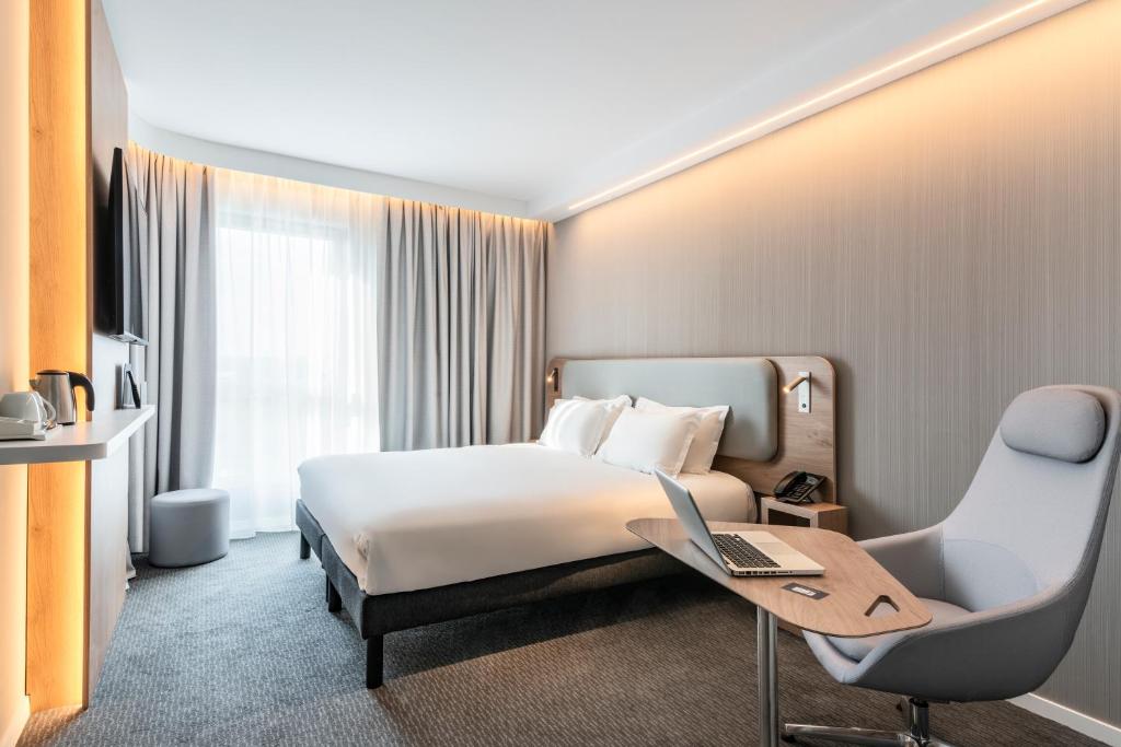 Hotel room for group accommodation, with grey carpet flooring, featuring a double bed with a light grey upholstered headboard and white bedding in the centre, a wooden desk with an open laptop and a grey ergonomic chair in the foreground on the right, a flat-screen TV and kettle on a white shelf set into a warm-lit wood alcove on the left, and floor-to-ceiling grey curtains in front of the window in the background.