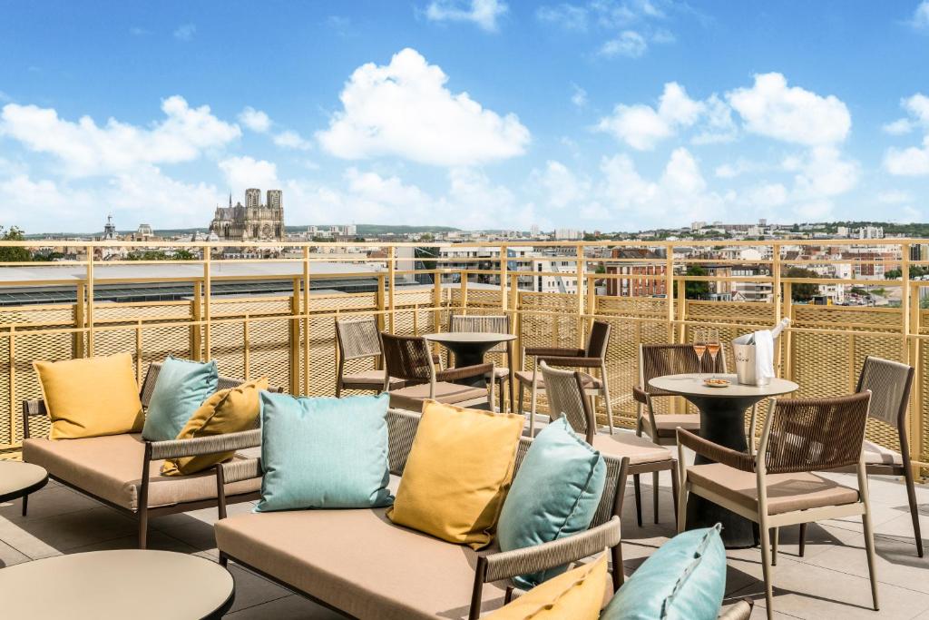 Hotel rooftop terrace with rattan sofas and armchairs with yellow and teal cushions, small round tables with champagne glasses, a golden mesh railing, and a panoramic view of the city with a Gothic cathedral visible on the left under a blue sky, for a rooftop afterwork.