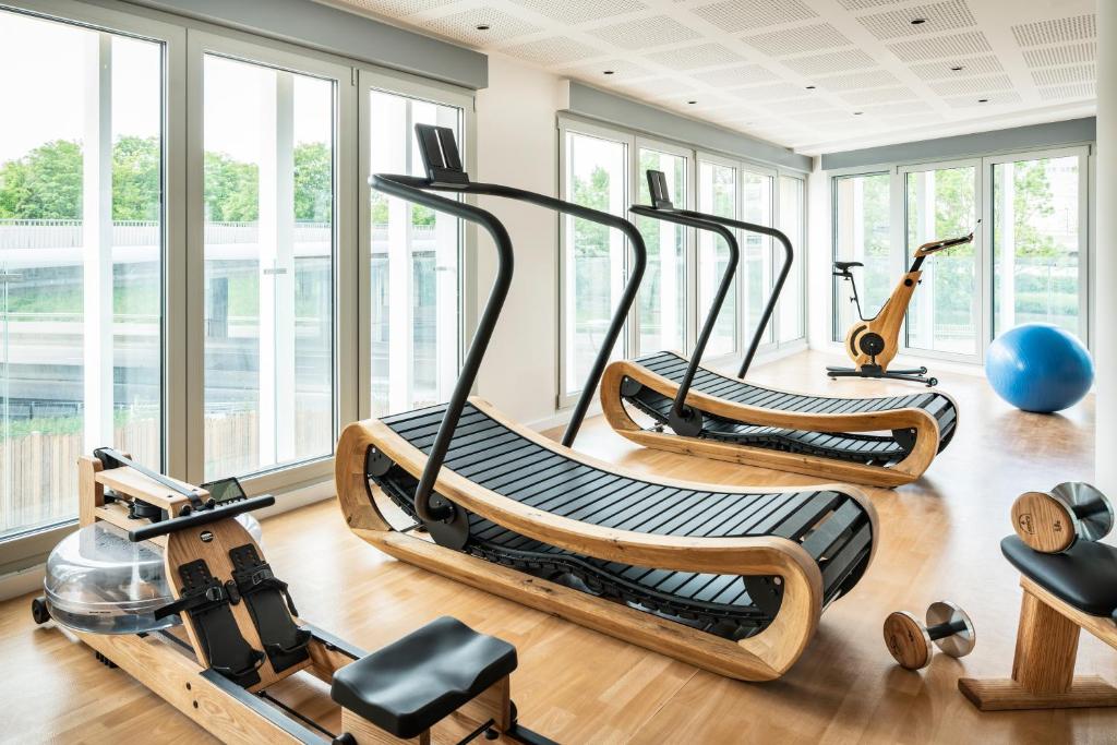 Hotel fitness room for a wellness seminar, with light wood flooring and large bay windows overlooking greenery on two sides, featuring two curved wooden-framed treadmills in the centre, a rowing machine on the left, a wooden spinning bike and a blue fitness ball in the background on the right, and wooden dumbbells on the floor in the foreground.