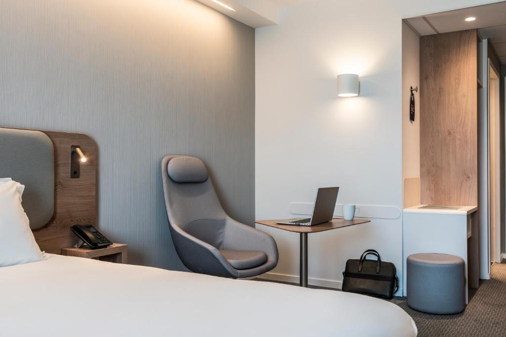 Hotel room with grey carpet and light walls, featuring a double bed with a wooden headboard and reading sconce on the left, a grey ergonomic armchair and a small wooden desk with a laptop in the centre, a round grey pouf and a light wood wardrobe area on the right.