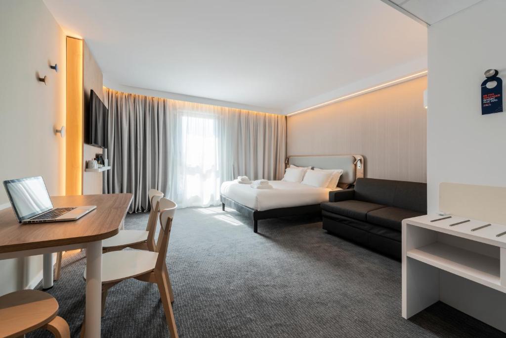 Spacious hotel room with grey carpet and warm LED lighting, featuring a round wooden table with chairs and a laptop on the left, a double bed with a padded headboard and a dark sofa in the centre, a wall-mounted television and grey curtains in the background, for group accommodation.