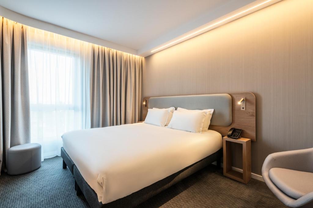 Hotel double bed room with grey carpet and warm LED ceiling lighting, featuring a double bed with a grey padded headboard framed in light wood with reading sconces, a wooden bedside table with a telephone, a large window with beige curtains on the left, a grey round pouf and a white armchair.
