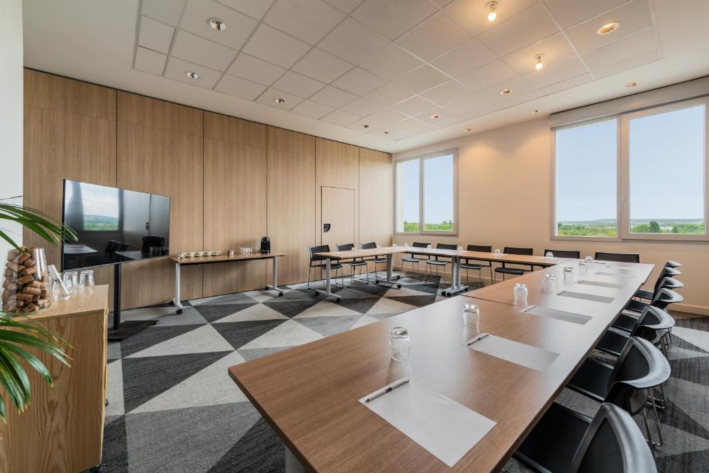 Room rental space arranged in a U-shape configuration, featuring tables equipped with notepads, pens, glasses, and water bottles surrounding the center area, black chairs distributed throughout, large windows on the right overlooking the landscape, and a presentation screen positioned on the left; wood-paneled walls and geometric carpeting create a bright, professional setting.