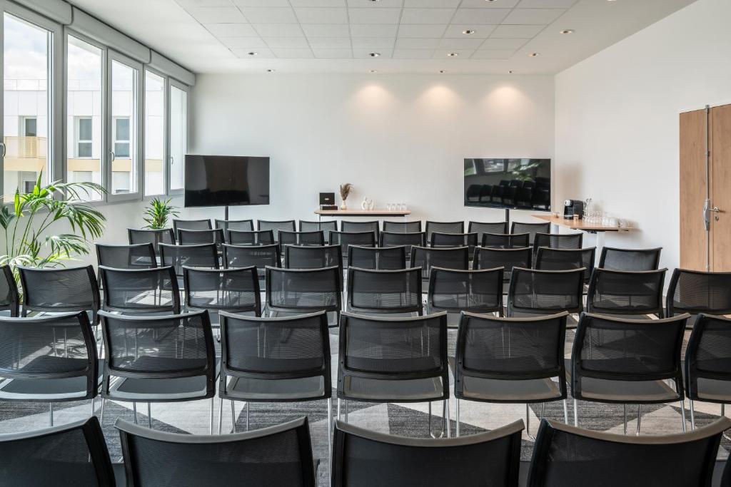 Training room rental space with rows of black chairs arranged in theater style across the foreground and center, featuring large windows along the left wall, presentation screens positioned at the front, and refreshment stations set up against the rear wall; bright white walls and recessed ceiling lighting create a modern professional environment.