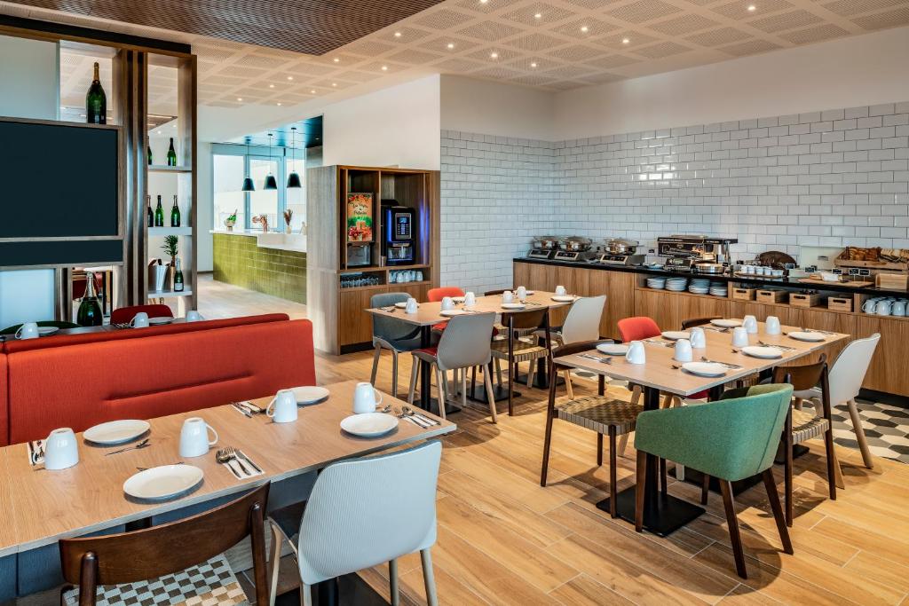 Hotel restaurant with on-site dining, with light wood flooring and a checkered tile area in the foreground, wooden tables set with white plates and cups, multicoloured chairs in grey, red and green, a red banquette on the left, a buffet counter along the white subway-tiled wall on the right, and a bar area with dark pendant lights and a green-tiled column visible in the background.