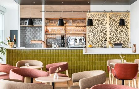 Hotel bar with a green ribbed tile counter and white marble top, featuring an espresso machine, backlit glass shelving with bottles and glasses, decorative golden laser-cut panels on the right, black pendant lights overhead, and pink and beige rounded armchairs with small round tables in the foreground, for an afterwork drink.