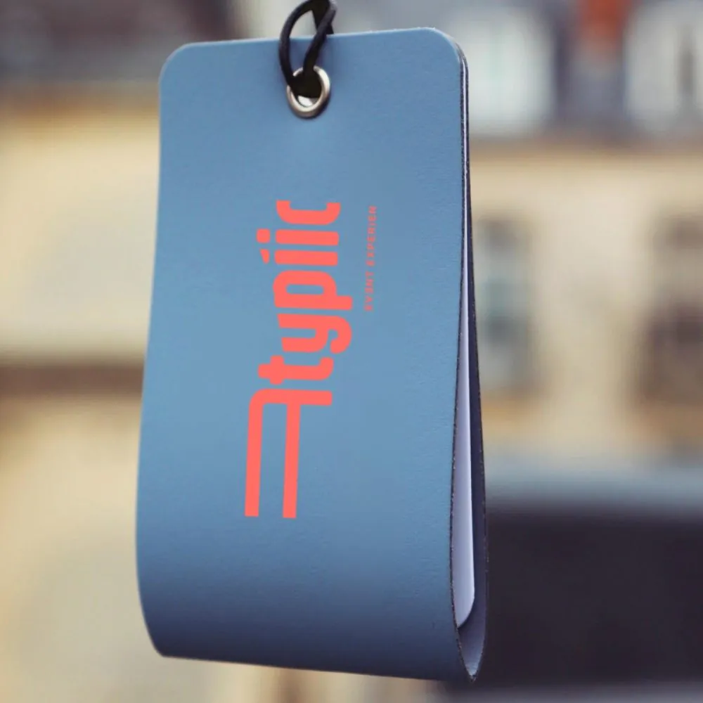 Gros plan sur une étiquette à bagage bleue avec le logo "Atypiic" en rouge, suspendue à un anneau métallique noir sur fond de façade haussmannienne floue en arrière-plan.