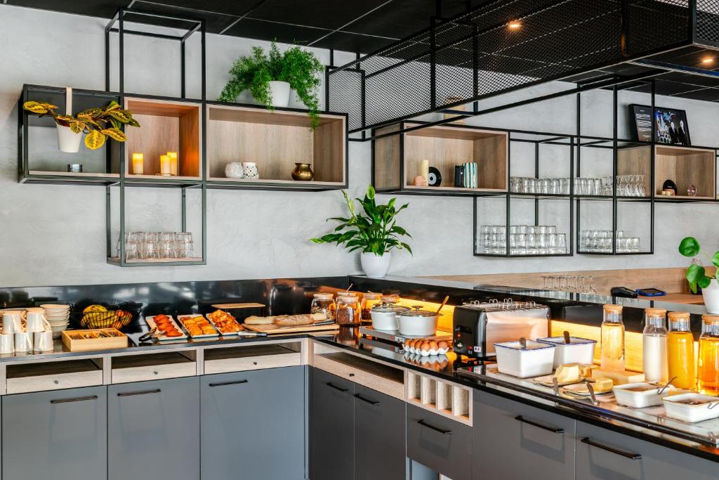 Hotel breakfast buffet with grey cabinetry and a black countertop, featuring pastries, eggs, cold cuts, juice bottles, white bowls and condiments along a lit L-shaped counter, topped by open black metal and wood shelving units with glasses, plants, candles and decorative objects, under a black metal mesh ceiling.